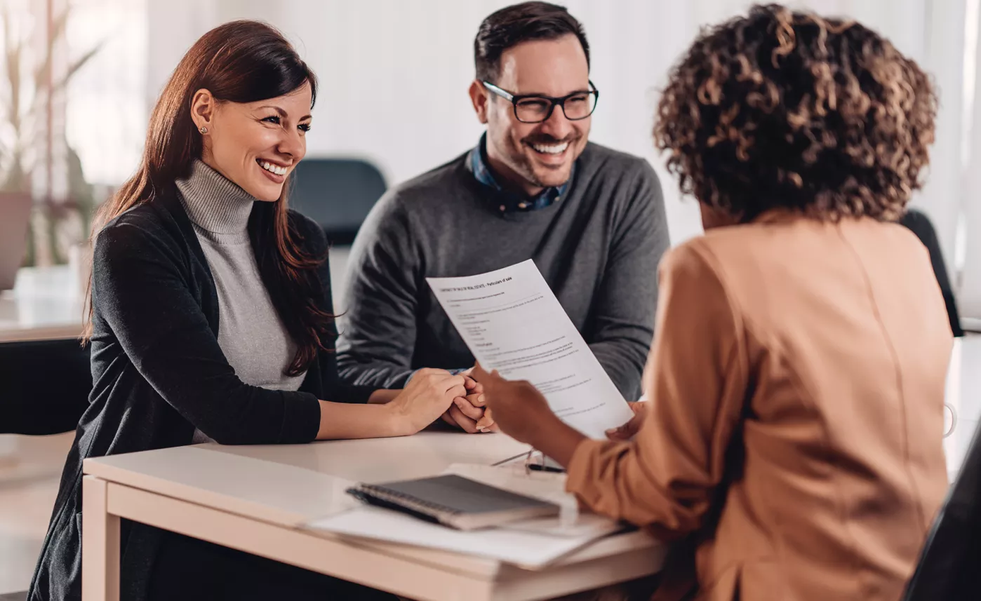  A smiling couple, sitting at a table reviewing documents with a financial advisor.
