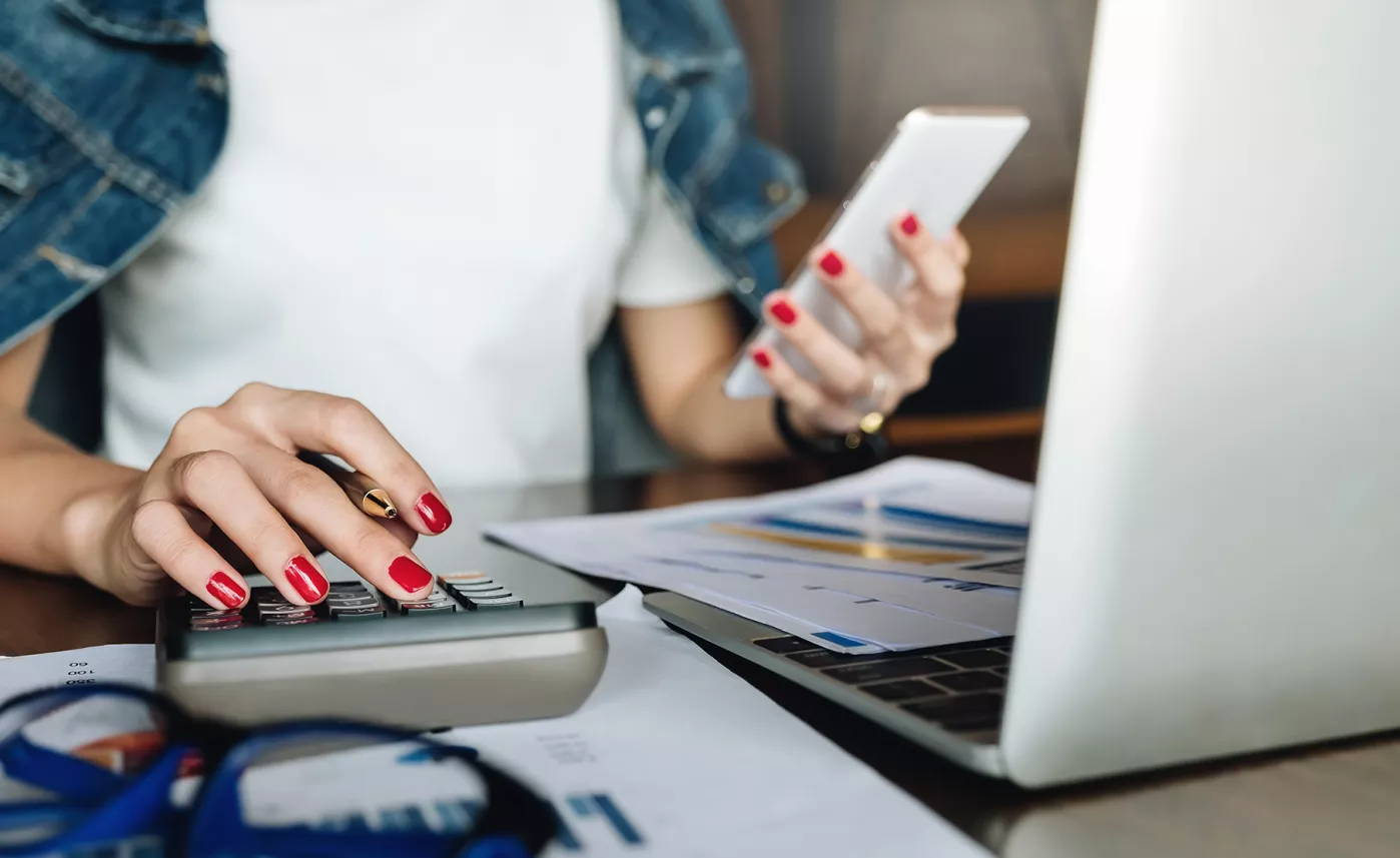  A woman reads a mobile phone and types figures into a calculator, an open laptop and financial documents in front of her.
