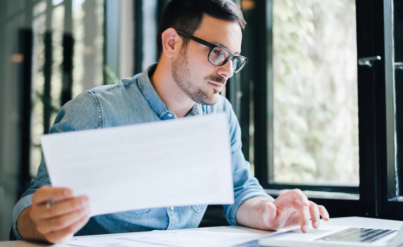  A man reads through sheets of paper while working on his laptop in an office.
