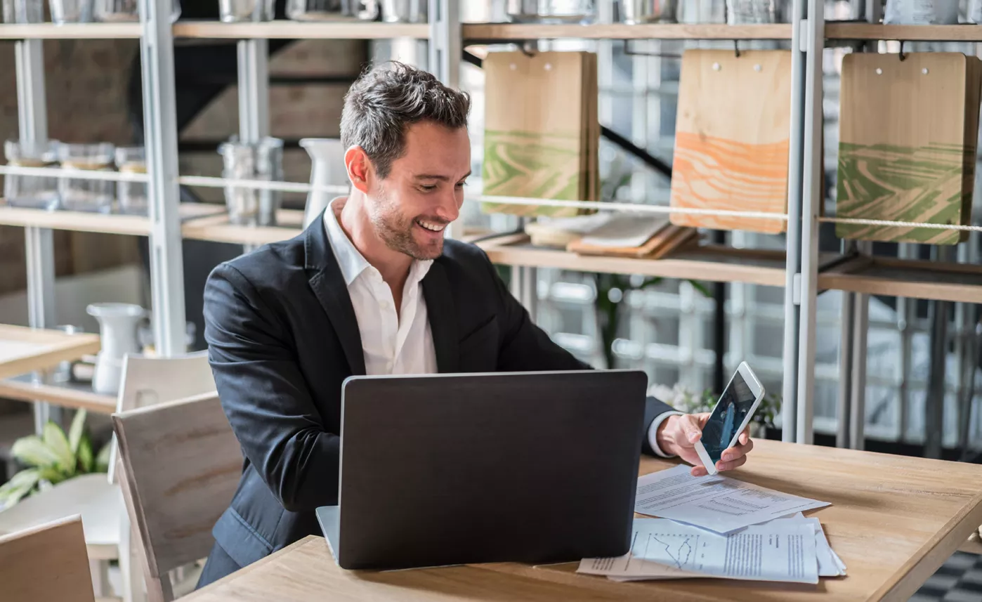 A man reviews financial documents on his laptop and phone at his small business.
