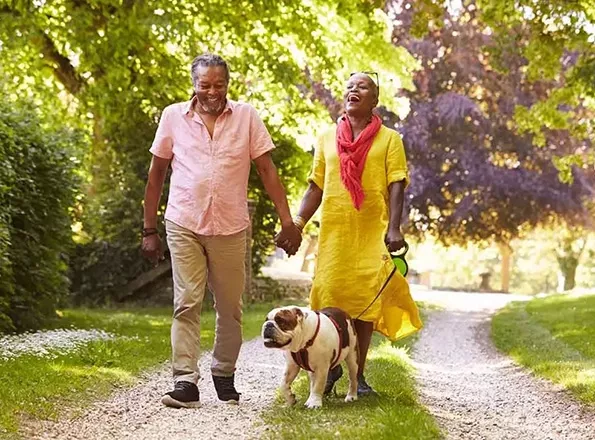  An older couple walking their dog in the park, engaged in lighthearted conversation and laughter.
