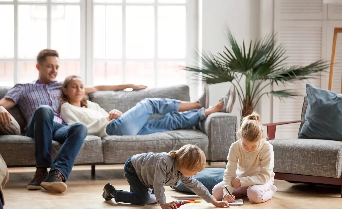  Parents sitting on the couch in the family room. They are smiling and watching over their children while they draw on their coloring books.
