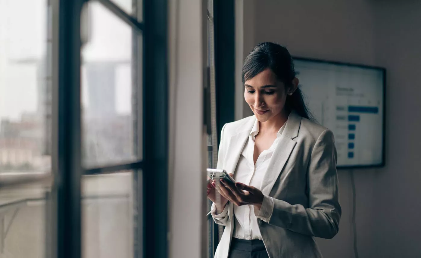  A professionally dressed person, standing by a window, looking at their mobile phone.

