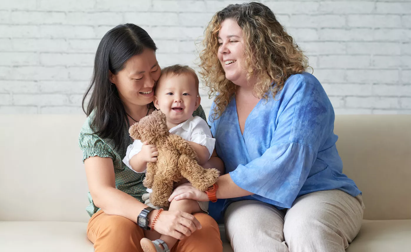  Two people smiling, sitting on a couch with a baby holding a stuffed animal.
