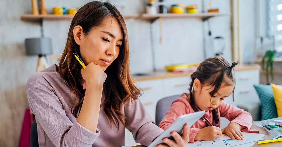  Parent and young child sitting at table. The parent is looking at their phone in contemplation while the child colours.
