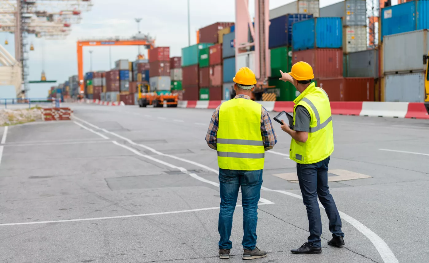  Two men in yellow vests working in a shipping yard.
