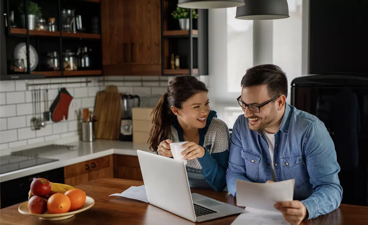  A couple sitting and laughing together at a kitchen counter looking at a laptop and paperwork.
