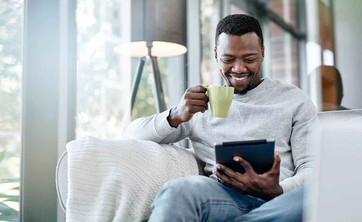  A man drinks from a coffee mug while smiling and using a tablet device.
