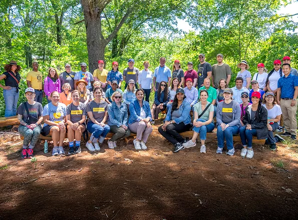  A large group of people posing for a photo in a wooded area, with trees and greenery in the background. They are wearing casual clothing and hats.
