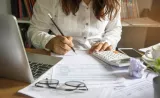 A woman reviews and calculates tax information in her home office, with documents spread out on her desk.