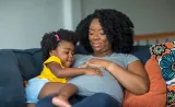 A mother lies on the couch with her young daughter, each with a hand on the mother's pregnant stomach.