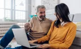 A couple smiles at each other while they do their taxes together in the living room.