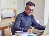 A man checks his finances in front of his laptop at his kitchen table, and takes notes in a logbook.