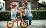 A parent putting a helmet on their young child who is sitting on a bike.