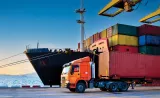 A cargo ship docked at a port, with colorful shipping containers stacked on it and an orange truck transporting a container nearby.